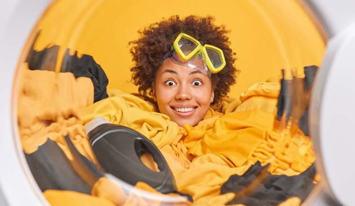 Happy pleased housewife smiles broadly enjoys washing loads washer with laundry uses liquid powder wears snorkeling glasses on forehead busy doing housework.