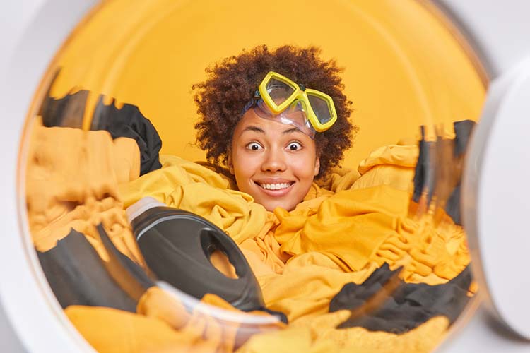 Happy pleased housewife smiles broadly enjoys washing loads washer with laundry uses liquid powder wears snorkeling glasses on forehead busy doing housework.